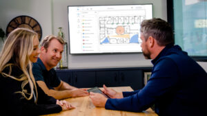 Three people are engaged in a discussion at a table, with a digital map displayed on a screen behind them.
