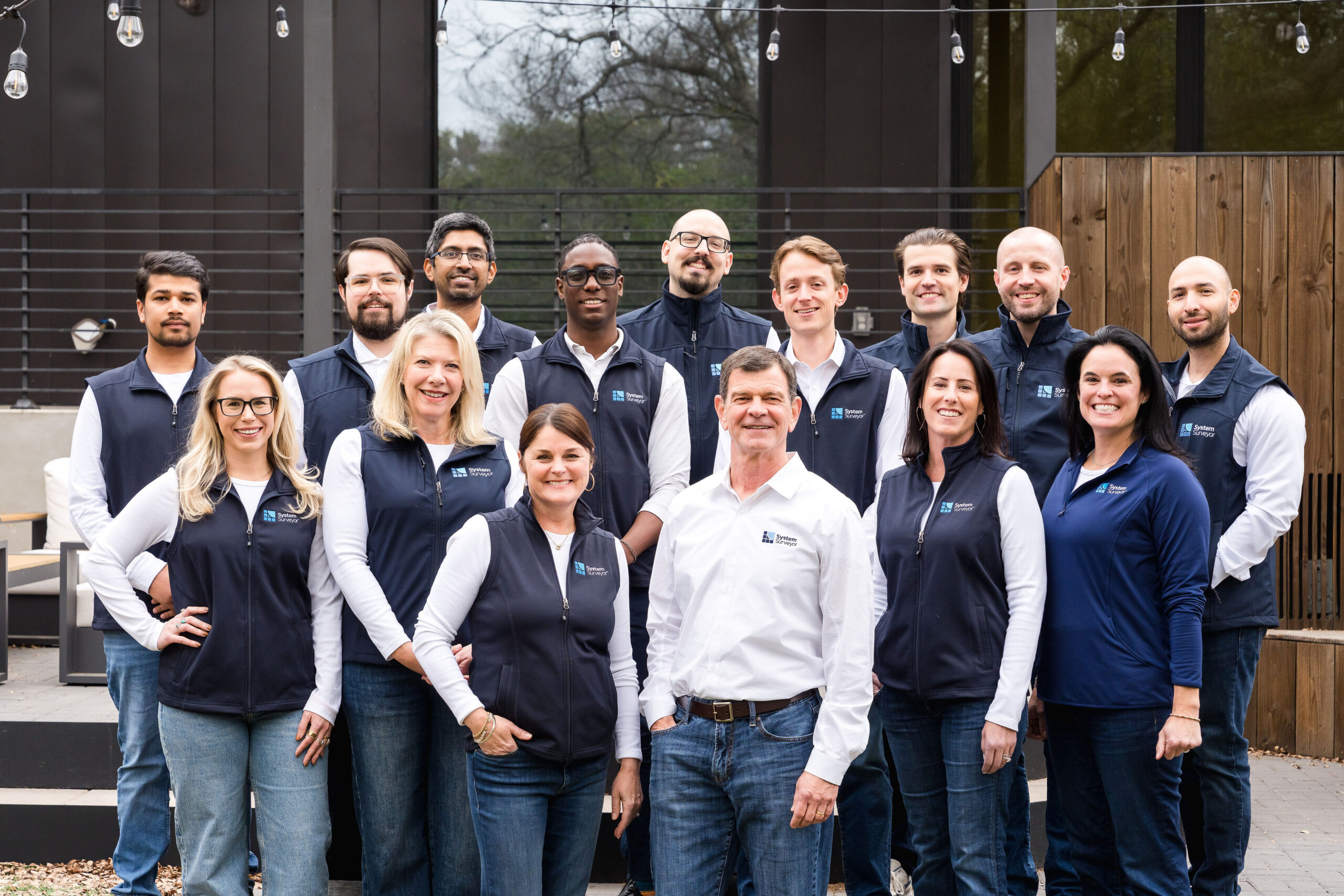 A diverse group of 15 people poses together outdoors, wearing matching vests and casual attire, in a modern setting.