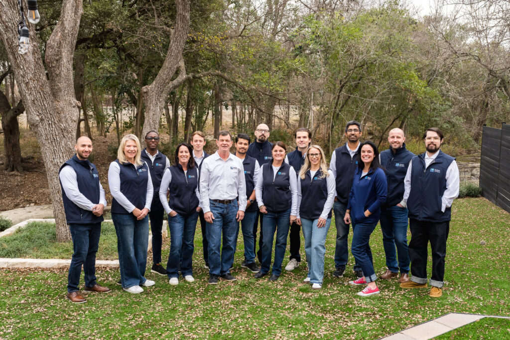 A group photo of 15 people in casual clothing, standing outdoors in a grassy area surrounded by trees.