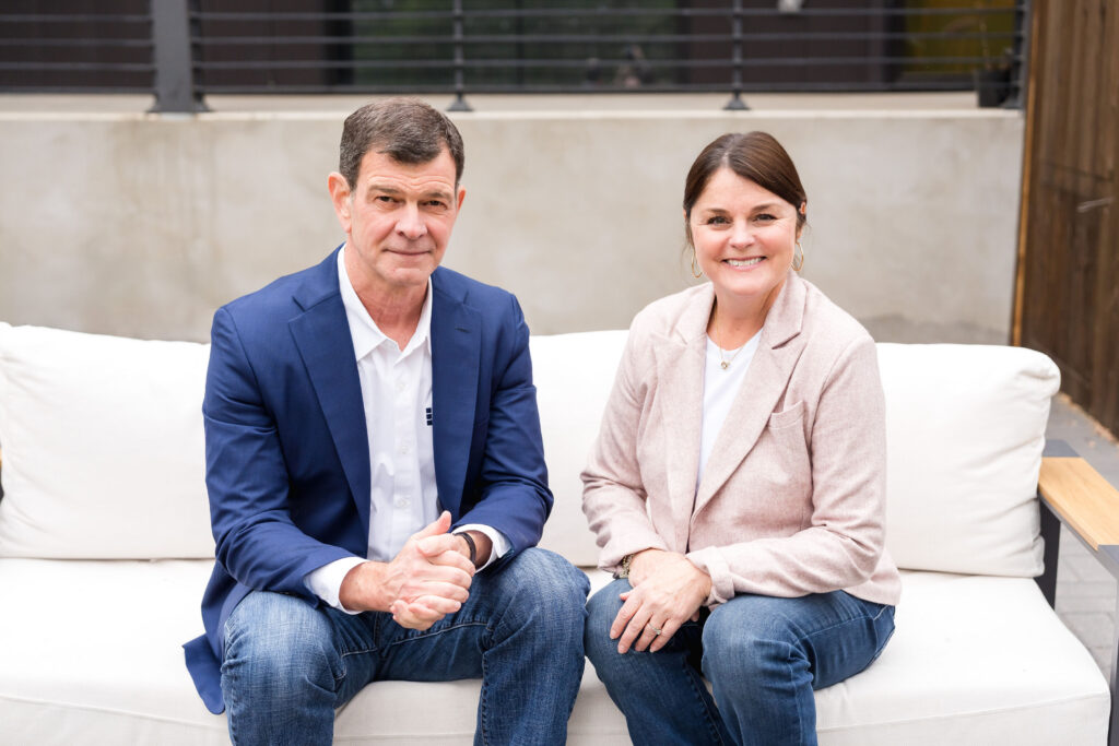 A man in a blazer and a woman in a light jacket sit together on a sofa, smiling in a casual outdoor setting.
