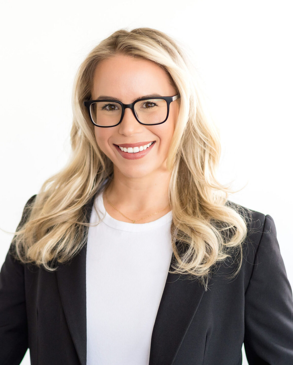 A smiling woman with blonde hair and glasses wears a black blazer and white top, set against a bright background.